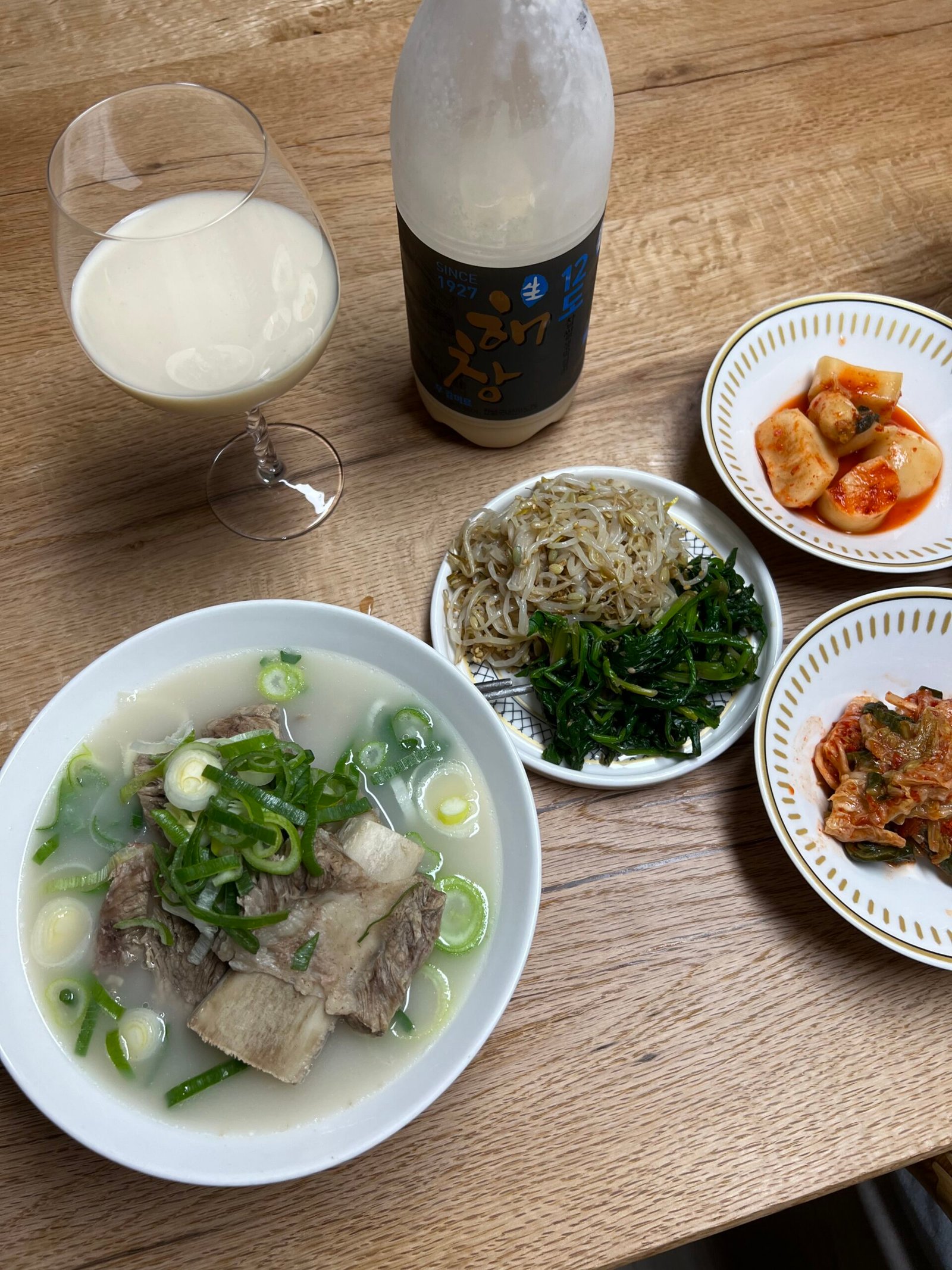 A glass of haechang makgeolli poured beside a full Korean meal with soup, greens, bean sprouts and kimchi on a wooden table