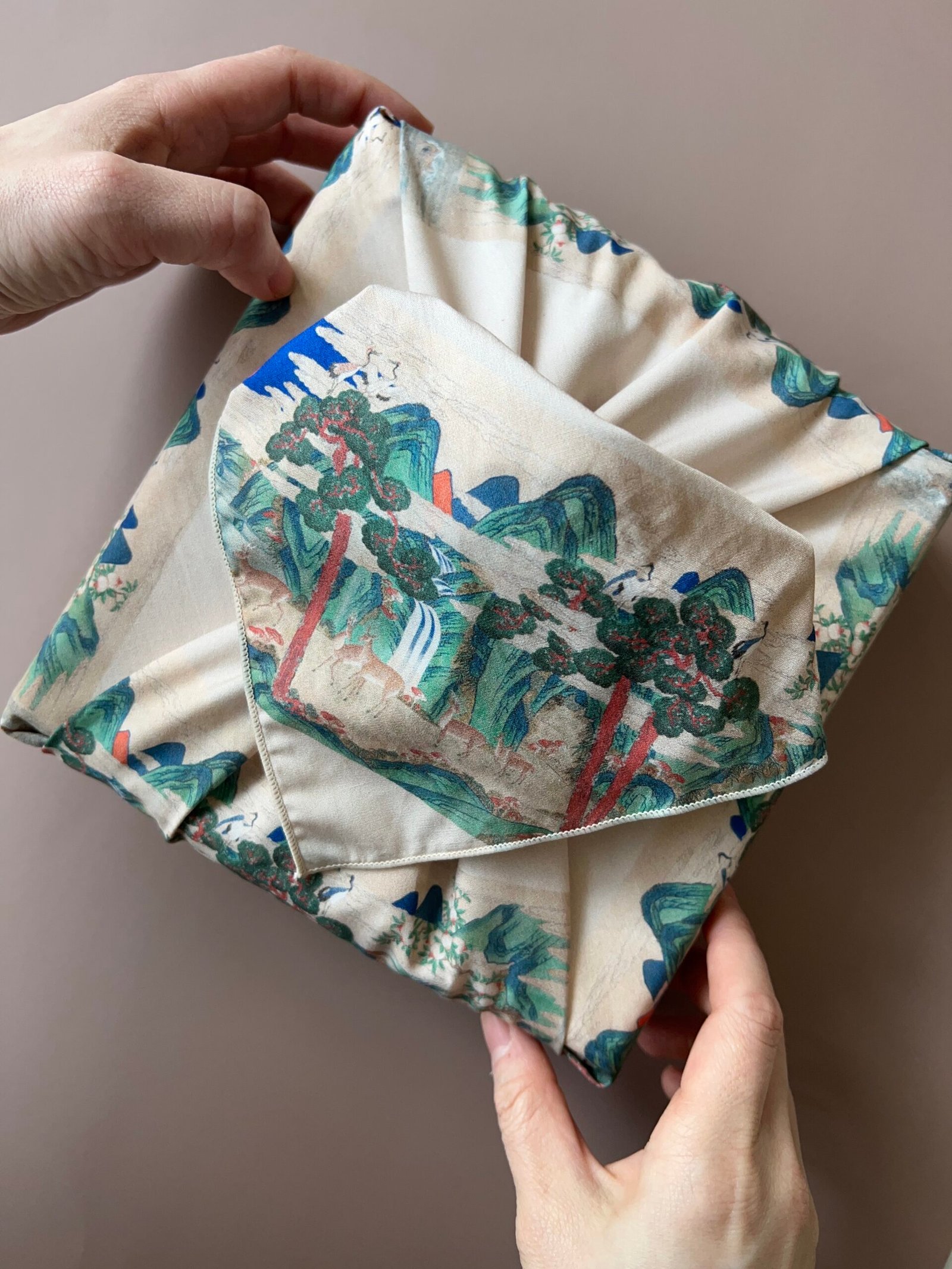 Hands folding a bojagi printed with Sipjangsaeng motifs of pine, cranes, and deer